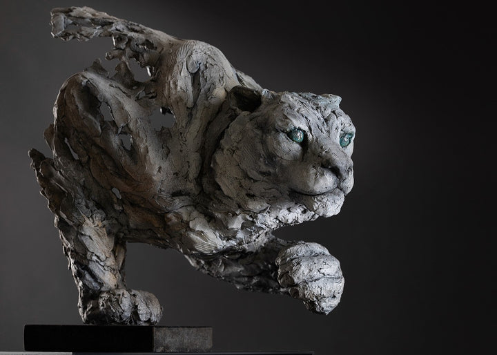 Sculpture of a stalking snow leopard by Lucy d’Auvergne, created in cast slate with raw earth pigments, showing a powerful crouched pose with textured grey and earthy tones, green eyes, and mounted on a granite base.