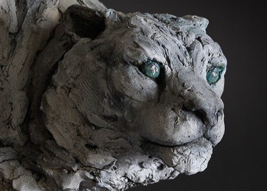 Sculpture of a stalking snow leopard by Lucy d’Auvergne, created in cast slate with raw earth pigments, showing a powerful crouched pose with textured grey and earthy tones, green eyes, and mounted on a granite base.
