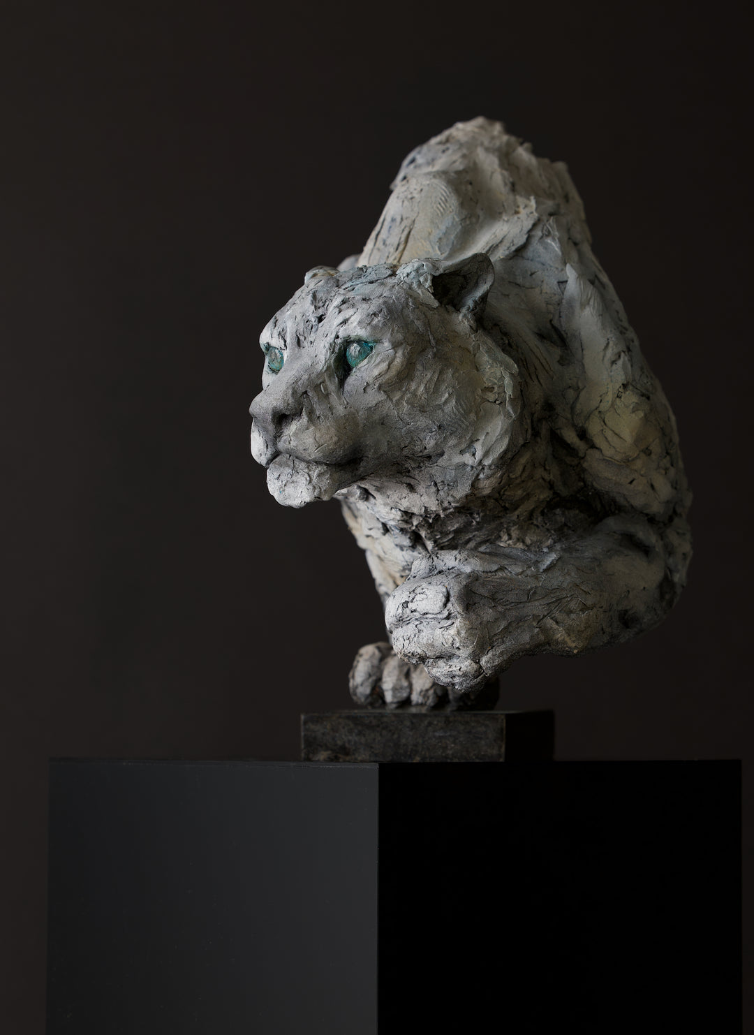 Sculpture of a stalking snow leopard by Lucy d’Auvergne, created in cast slate with raw earth pigments, showing a powerful crouched pose with textured grey and earthy tones, green eyes, and mounted on a granite base.