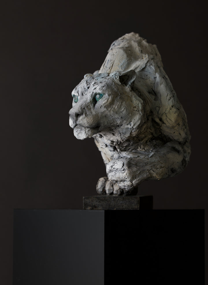 Sculpture of a stalking snow leopard by Lucy d’Auvergne, created in cast slate with raw earth pigments, showing a powerful crouched pose with textured grey and earthy tones, green eyes, and mounted on a granite base.