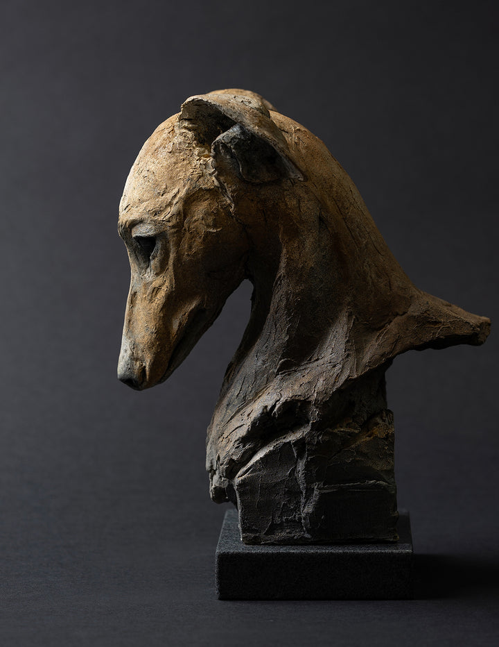 Sculpture of a contemplative dog’s head by Lucy d’Auvergne, cast in slate with raw earth pigments creating warm ochre and umber tones with deep charcoal shadows, mounted on a polished granite base.