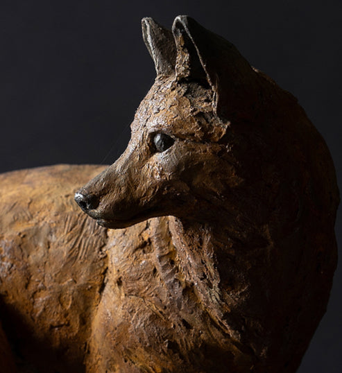 Sculpture of a young fox by Lucy d’Auvergne, cast in cold cast iron with warm earthy brown patina, shown standing alert and turning its head, mounted on a natural oak plinth.