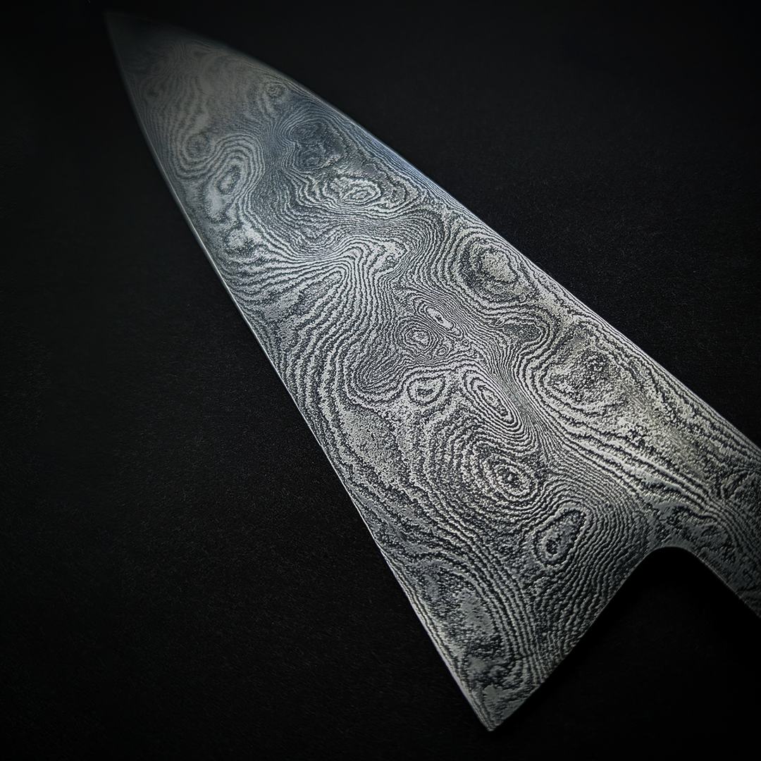 Close-up and full view of a handmade Damascus steel chef’s knife by Beggs Forgeworks, showcasing the intricate layered grain of the forged blade. The unique patterning of the steel, created through repeated folding and etching, is paired with a dark, faceted wooden handle—highlighting the craftsmanship and balance of the blade against a matte black background.