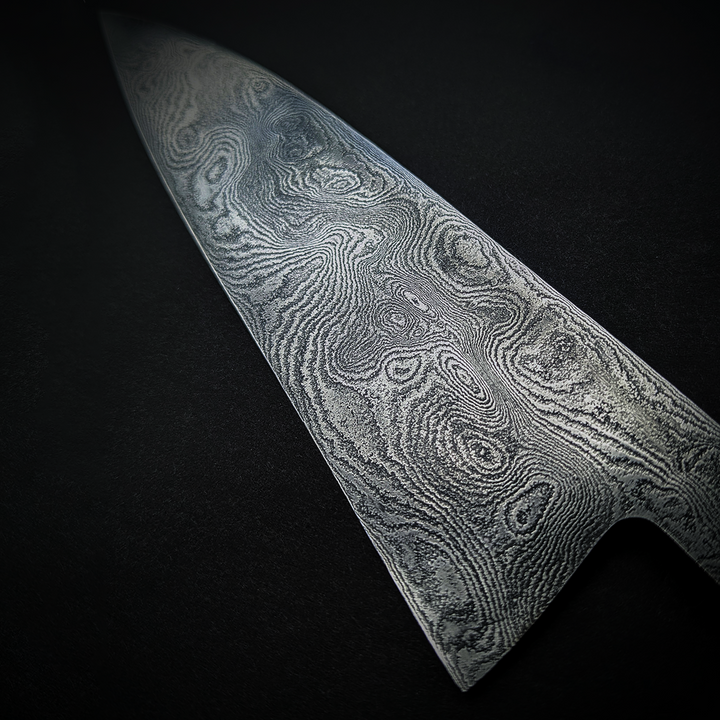 Close-up and full view of a handmade Damascus steel chef’s knife by Beggs Forgeworks, showcasing the intricate layered grain of the forged blade. The unique patterning of the steel, created through repeated folding and etching, is paired with a dark, faceted wooden handle—highlighting the craftsmanship and balance of the blade against a matte black background.