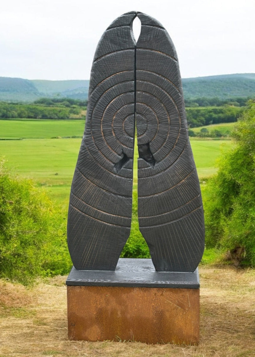 Glen Farrelly sculpture First Breath Again, a 125cm tall one-off carved from reclaimed unwanted oak. The form features two upright sections with a central split, radiating circular carvings echoing tree rings and energy pulses. Charred blackened oak reveals warm golden-brown undertones, symbolising renewal, sustainability, and the power of nature reclaimed. A striking contemporary work for both gardens and interior display, balancing raw presence with contemplative form.