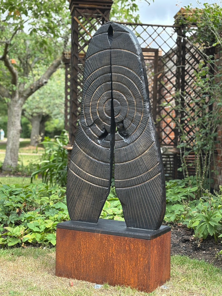 Glen Farrelly sculpture First Breath Again, a 125cm tall one-off carved from reclaimed unwanted oak. The form features two upright sections with a central split, radiating circular carvings echoing tree rings and energy pulses. Charred blackened oak reveals warm golden-brown undertones, symbolising renewal, sustainability, and the power of nature reclaimed. A striking contemporary work for both gardens and interior display, balancing raw presence with contemplative form.