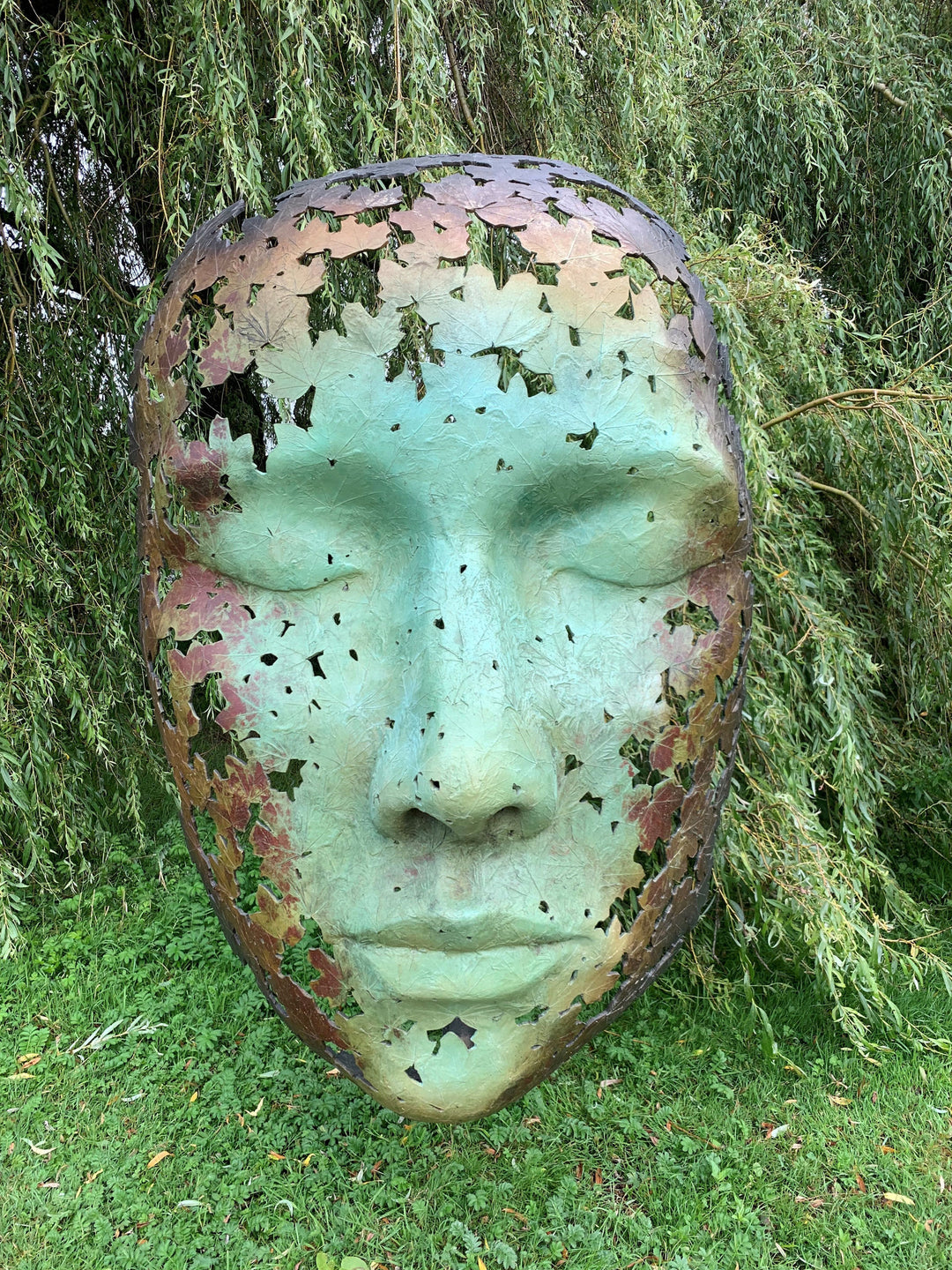 Simon Gudgeon bronze sculpture Leaf Spirit featuring a serene monumental face formed with organic leaf patterns displayed within a landscaped garden and woodland setting.