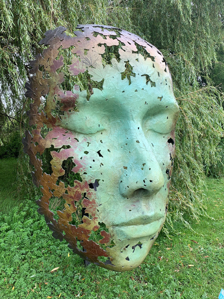 Simon Gudgeon bronze sculpture Leaf Spirit featuring a serene monumental face formed with organic leaf patterns displayed within a landscaped garden and woodland setting.