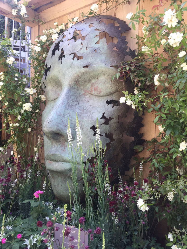 Simon Gudgeon bronze sculpture Leaf Spirit featuring a serene monumental face formed with organic leaf patterns displayed within a landscaped garden and woodland setting.