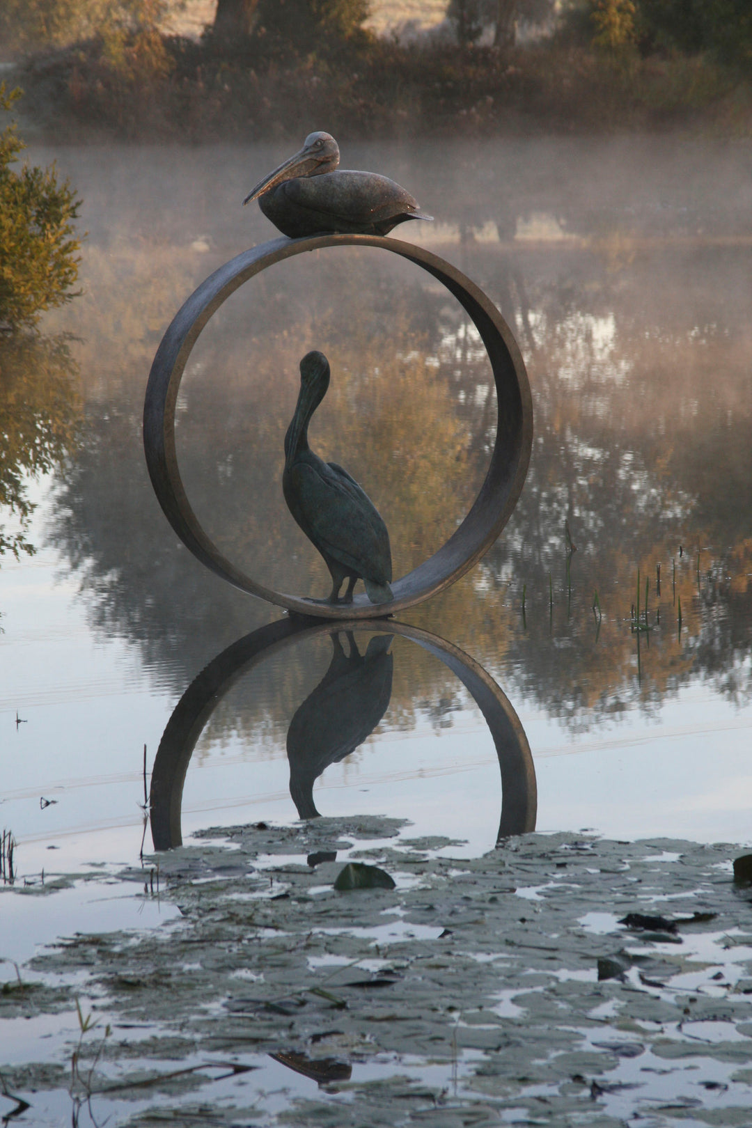 Simon Gudgeon bronze sculpture Pelicans featuring two elegant stylised pelican forms positioned around a circular contemporary bronze structure within a landscaped outdoor setting.