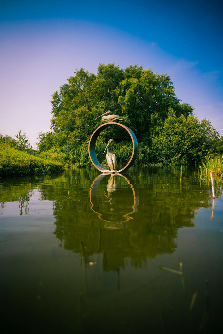 Simon Gudgeon bronze sculpture Pelicans featuring two elegant stylised pelican forms positioned around a circular contemporary bronze structure within a landscaped outdoor setting.