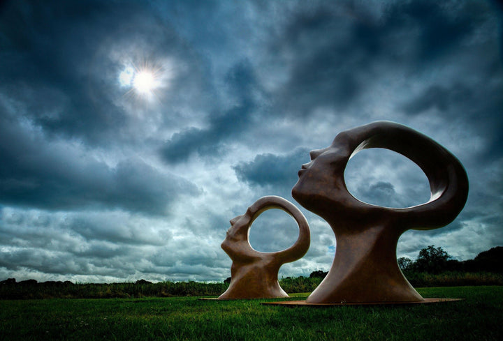 Simon Gudgeon bronze sculpture Search for Enlightenment featuring monumental interlocking abstract human forms with circular openings displayed within a landscaped outdoor setting.