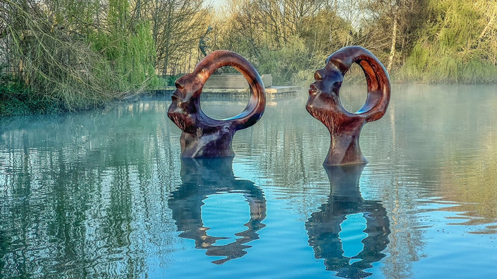 Simon Gudgeon bronze sculpture Search for Enlightenment featuring monumental interlocking abstract human forms with circular openings displayed within a landscaped outdoor setting.