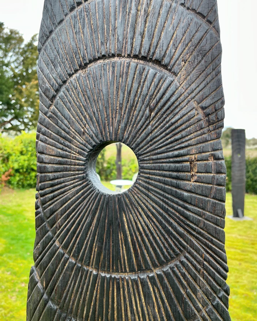 Glen Farrelly sculpture Gobaith, a 205cm tall standing form carved from unwanted oak using the Yakisugi technique, inspired by ancient standing stones. Featuring radiating carved ring-mark patterns and a central circular aperture, the charred blackened surface reveals warm natural oak tones beneath, creating a striking presence of resilience, hope, and renewal in both garden and public spaces.
