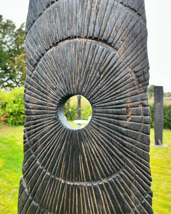 Glen Farrelly sculpture Gobaith, a 205cm tall standing form carved from unwanted oak using the Yakisugi technique, inspired by ancient standing stones. Featuring radiating carved ring-mark patterns and a central circular aperture, the charred blackened surface reveals warm natural oak tones beneath, creating a striking presence of resilience, hope, and renewal in both garden and public spaces.