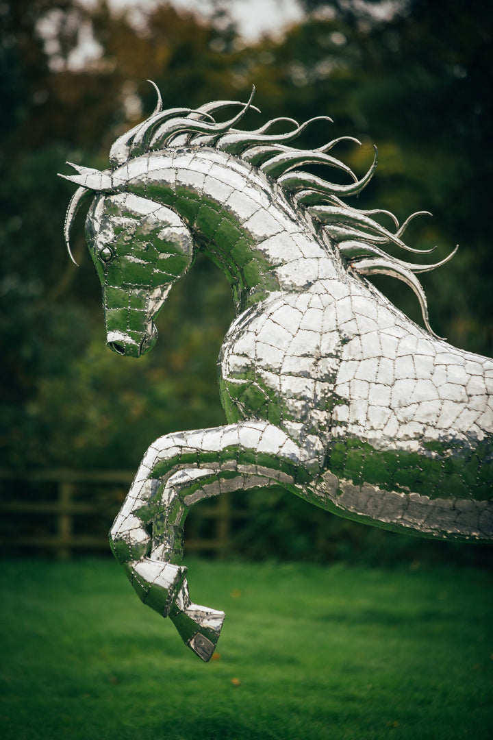 ‘Leaping Horse’ by Michael Turner, a unique, one-of-one stainless steel equestrian sculpture depicting a magnificent life-size horse leaping forward with its front legs raised and hind legs extended. Handcrafted from marine-grade stainless steel, the sculpture features intricately welded metallic plates, a powerful muscular body, flowing mane and sweeping tail. Its polished silver finish reflects the surrounding landscape, while the dynamic pose captures the strength, elegance and movement of a horse in ful