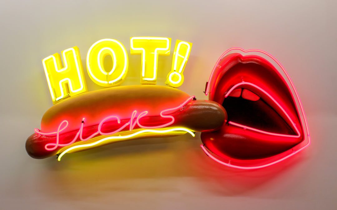 Playful neon wall art by Chris Bracey featuring a hot dog with the words “HOT Licks” in glowing yellow and pink neon, paired with a bold red neon mouth and lips design. Sculptural elements include a fabricated hot dog and 3D lips, combining humour and innuendo in classic Bracey style.