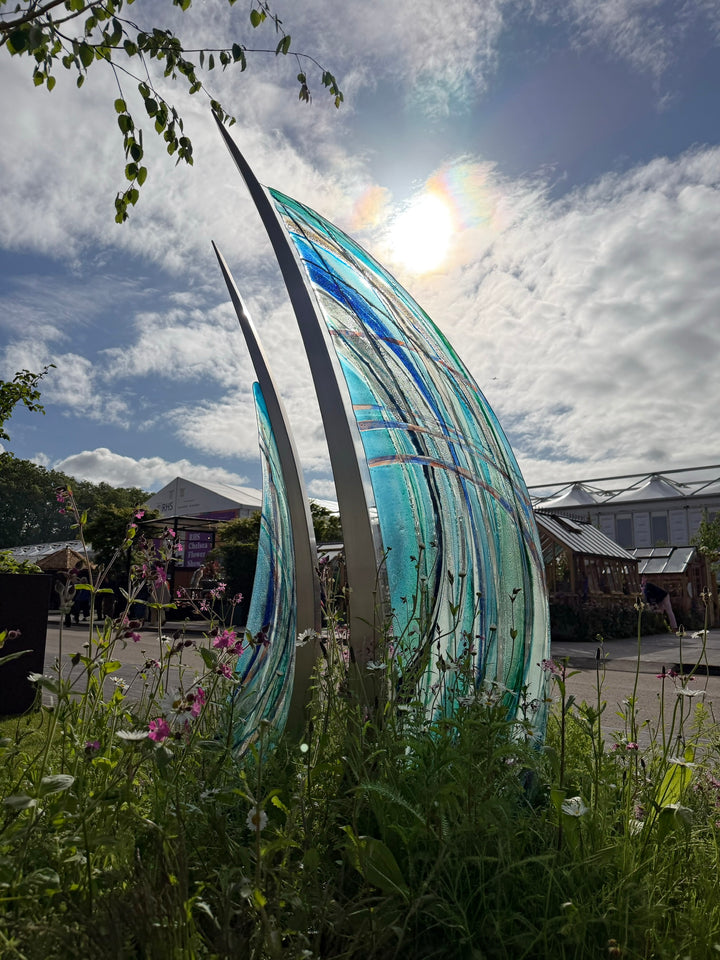 Carrie-Anne Funnell glass and stainless steel sculpture Sail Away, featuring two large curved sail-like forms with layered blue, turquoise and aqua glass framed in polished stainless steel, displayed within a landscaped outdoor garden setting.