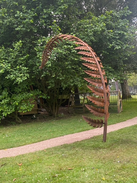 Towering corten steel fern sculptures form elegant archways within a lush garden landscape. Each sculptural frond, handcrafted from weathered steel, curves gracefully overhead and is designed as a water feature, releasing timed droplets into a pool below to create a tranquil, rhythmic ripple effect.
