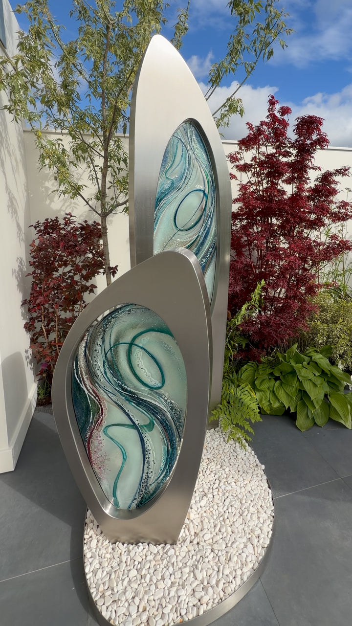 Double Apogee by Carrie-Anne Funnell, a large contemporary outdoor sculpture comprising two sweeping brushed stainless-steel forms inset with layered turquoise, blue and green kiln-formed glass.