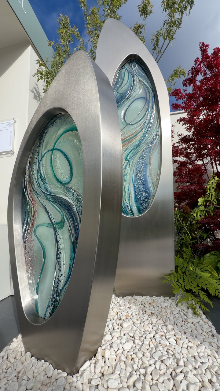 Double Apogee by Carrie-Anne Funnell, a large contemporary outdoor sculpture comprising two sweeping brushed stainless-steel forms inset with layered turquoise, blue and green kiln-formed glass.