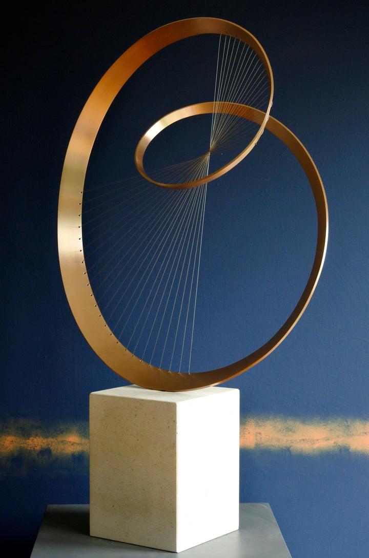 Fabricated bronze sculpture titled Infinity by Thomas Joynes, mounted on a Portland stone plinth. The work features two intersecting bronze curves connected by fine stainless steel micro wires, forming a minimal yet elegant spatial tension. The clean, modernist structure appears weightless and musical, its elliptical ribbons casting delicate linear shadows, evoking motion and balance. Photographed against a deep blue backdrop.