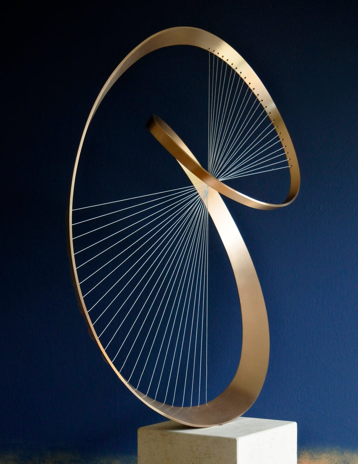 Fabricated bronze sculpture titled Infinity by Thomas Joynes, mounted on a Portland stone plinth. The work features two intersecting bronze curves connected by fine stainless steel micro wires, forming a minimal yet elegant spatial tension. The clean, modernist structure appears weightless and musical, its elliptical ribbons casting delicate linear shadows, evoking motion and balance. Photographed against a deep blue backdrop.