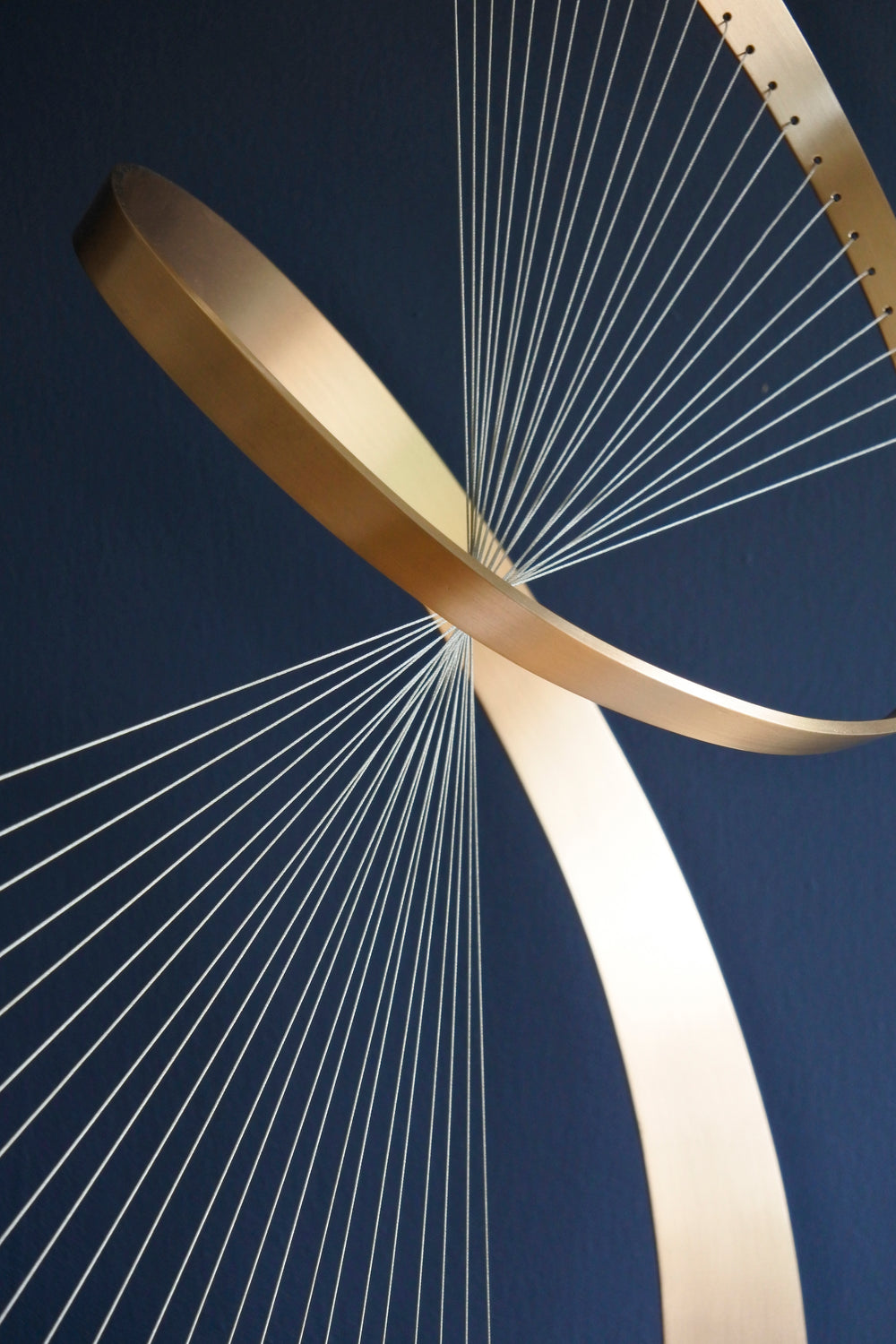 Fabricated bronze sculpture titled Infinity by Thomas Joynes, mounted on a Portland stone plinth. The work features two intersecting bronze curves connected by fine stainless steel micro wires, forming a minimal yet elegant spatial tension. The clean, modernist structure appears weightless and musical, its elliptical ribbons casting delicate linear shadows, evoking motion and balance. Photographed against a deep blue backdrop.