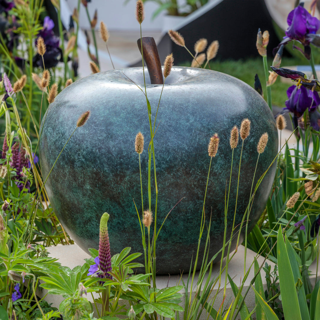 Blue-green patinated bronze apple sculpture by James Parker, positioned on a natural stone boulder within a landscaped garden setting.