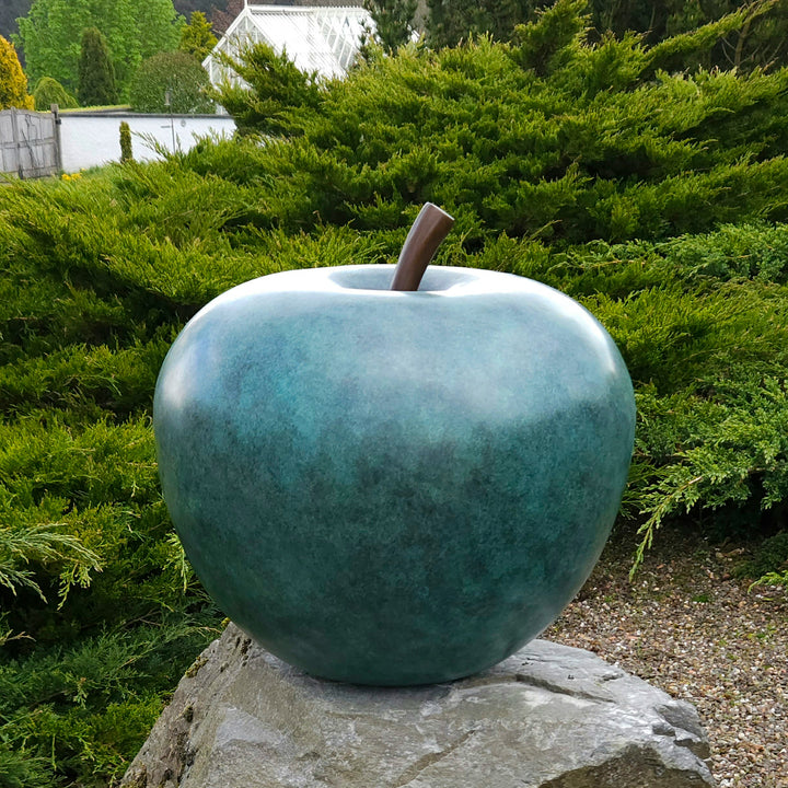 Blue-green patinated bronze apple sculpture by James Parker, positioned on a natural stone boulder within a landscaped garden setting.