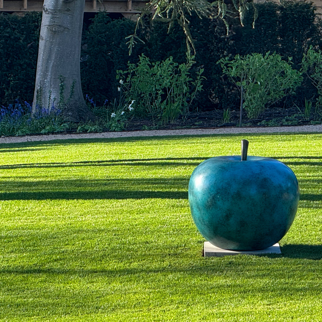 Blue-green patinated bronze apple sculpture by James Parker, positioned on a natural stone boulder within a landscaped garden setting.