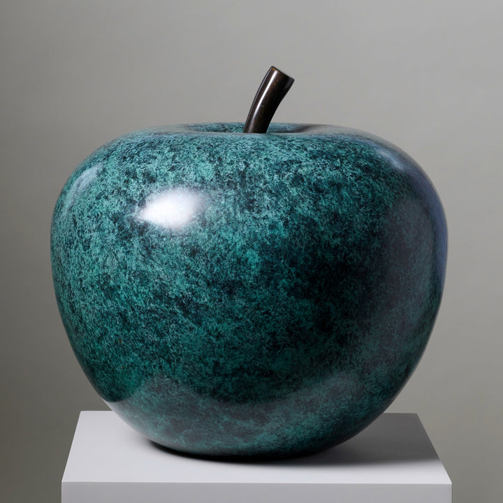 Blue-green patinated bronze apple sculpture by James Parker, positioned on a natural stone boulder within a landscaped garden setting.