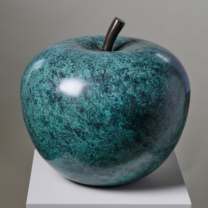 Blue-green patinated bronze apple sculpture by James Parker, positioned on a natural stone boulder within a landscaped garden setting.