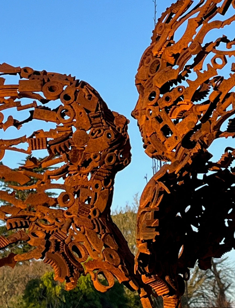 KISMET by Penny Hardy — a limited edition sculpture crafted from recycled metal, depicting two wind-blown figures leaning in to kiss, their bodies and hair formed from intricate mechanical parts and rusted fragments, mounted on a timber plinth in an open landscape.