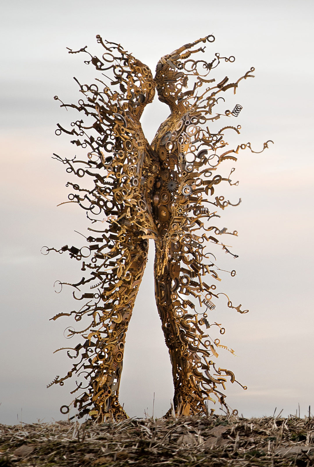 Blown Away by Penny Hardy — a striking figurative sculpture composed of welded reclaimed metal components, depicting two intertwined human forms dissolving into windblown fragments, captured against an open landscape and soft sky.