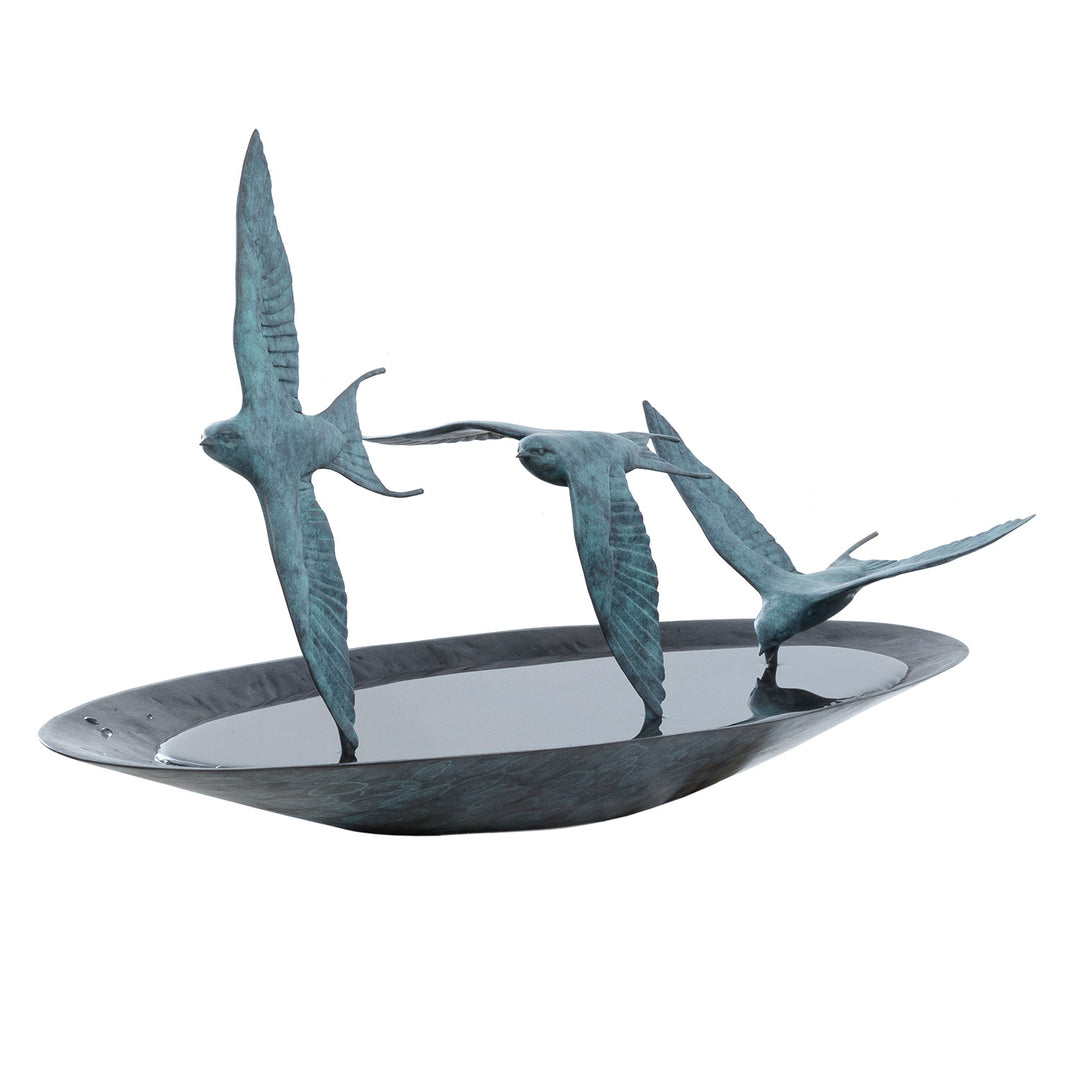 ‘Chasing Reflections’ by Pat Bull, a limited-edition contemporary bronze wildlife sculpture depicting three swallows flying low over a shallow, oval bronze water dish. The birds feature elegant outstretched wings, distinctive forked tails and finely textured feathers finished in turquoise and blue-green verdigris patina. Their wingtips appear to touch the smooth, reflective water surface, while the dark bronze dish creates a striking contrast with the lighter birds, capturing the movement and grace of swall
