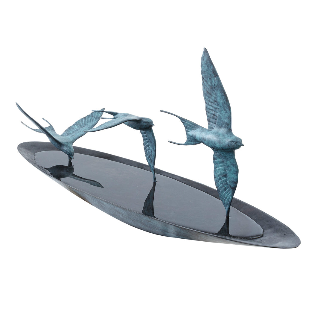 ‘Chasing Reflections’ by Pat Bull, a limited-edition contemporary bronze wildlife sculpture depicting three swallows flying low over a shallow, oval bronze water dish. The birds feature elegant outstretched wings, distinctive forked tails and finely textured feathers finished in turquoise and blue-green verdigris patina. Their wingtips appear to touch the smooth, reflective water surface, while the dark bronze dish creates a striking contrast with the lighter birds, capturing the movement and grace of swall