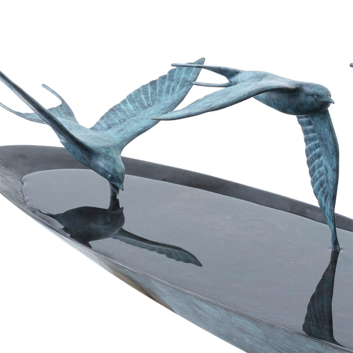 ‘Chasing Reflections’ by Pat Bull, a limited-edition contemporary bronze wildlife sculpture depicting three swallows flying low over a shallow, oval bronze water dish. The birds feature elegant outstretched wings, distinctive forked tails and finely textured feathers finished in turquoise and blue-green verdigris patina. Their wingtips appear to touch the smooth, reflective water surface, while the dark bronze dish creates a striking contrast with the lighter birds, capturing the movement and grace of swall