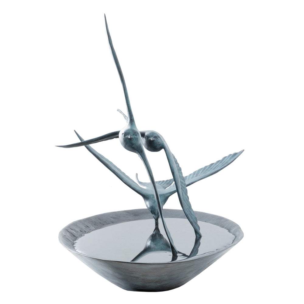 ‘Chasing Reflections’ by Pat Bull, a limited-edition contemporary bronze wildlife sculpture depicting three swallows flying low over a shallow, oval bronze water dish. The birds feature elegant outstretched wings, distinctive forked tails and finely textured feathers finished in turquoise and blue-green verdigris patina. Their wingtips appear to touch the smooth, reflective water surface, while the dark bronze dish creates a striking contrast with the lighter birds, capturing the movement and grace of swall