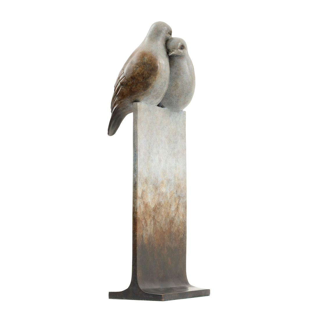 ‘Only Love’ by Pat Bull, a limited-edition contemporary bronze wildlife sculpture depicting two turtle doves perched closely together, their heads gently touching in an affectionate pose. The birds feature rounded bodies, delicate facial details and finely textured feathers in warm brown, soft grey, cream and pale bronze tones. Mounted on a tall rectangular bronze pedestal, the sculpture features a waxed, mottled patina fading from pale ivory and grey through earthy brown to dark charcoal and black, present