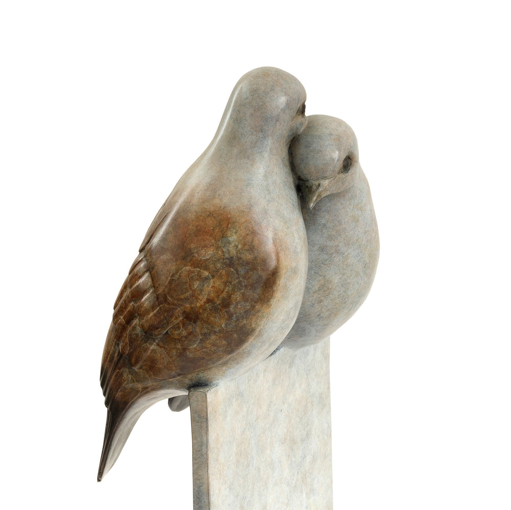 ‘Only Love’ by Pat Bull, a limited-edition contemporary bronze wildlife sculpture depicting two turtle doves perched closely together, their heads gently touching in an affectionate pose. The birds feature rounded bodies, delicate facial details and finely textured feathers in warm brown, soft grey, cream and pale bronze tones. Mounted on a tall rectangular bronze pedestal, the sculpture features a waxed, mottled patina fading from pale ivory and grey through earthy brown to dark charcoal and black, present