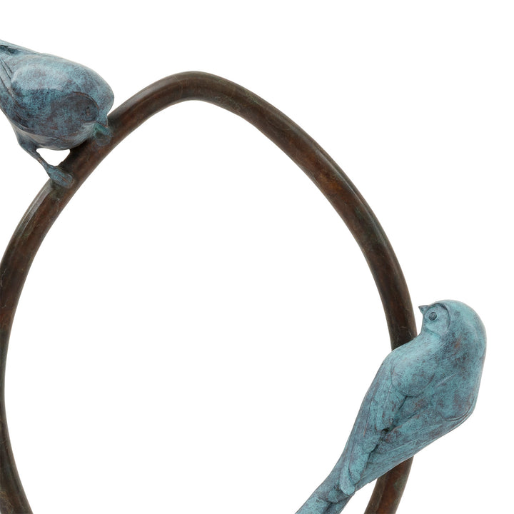 ‘Two’s Company’ by Pat Bull, a limited-edition contemporary bronze wildlife sculpture depicting two long-tailed tits perched on opposite sides of an upright circular bronze ring, facing towards one another. Each bird features a small rounded body, delicate beak, finely textured feathers and an elongated tail, finished in soft turquoise, teal and blue-green verdigris patina. The birds contrast with the dark brown and warm bronze tones of the open ring, which stands on a low oval bronze base against a white b