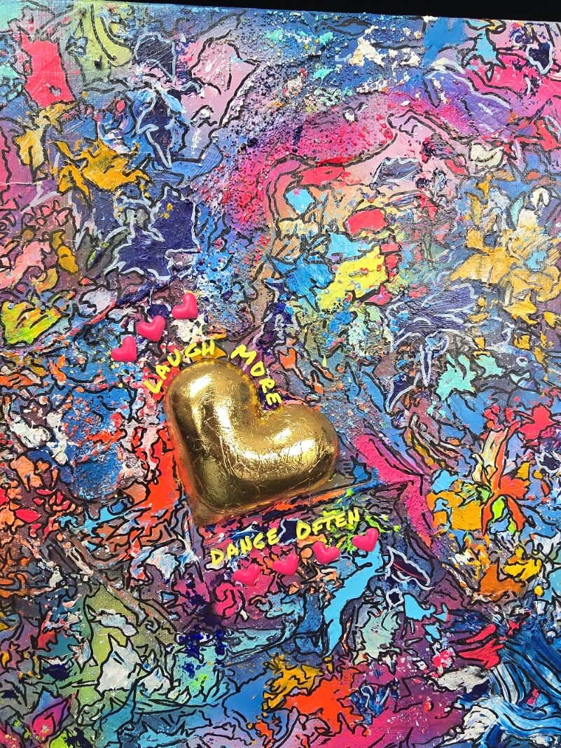 A vibrant abstract mixed-media painting bursting with swirling colours in pink, blue, yellow, orange and purple, with heavy texture and fluid forms. At the right side, a raised gold 3D heart sits prominently with text around it reading “Laugh More, Dance Often”. The artwork has an expressive, energetic surface with layered paint and resin details.
