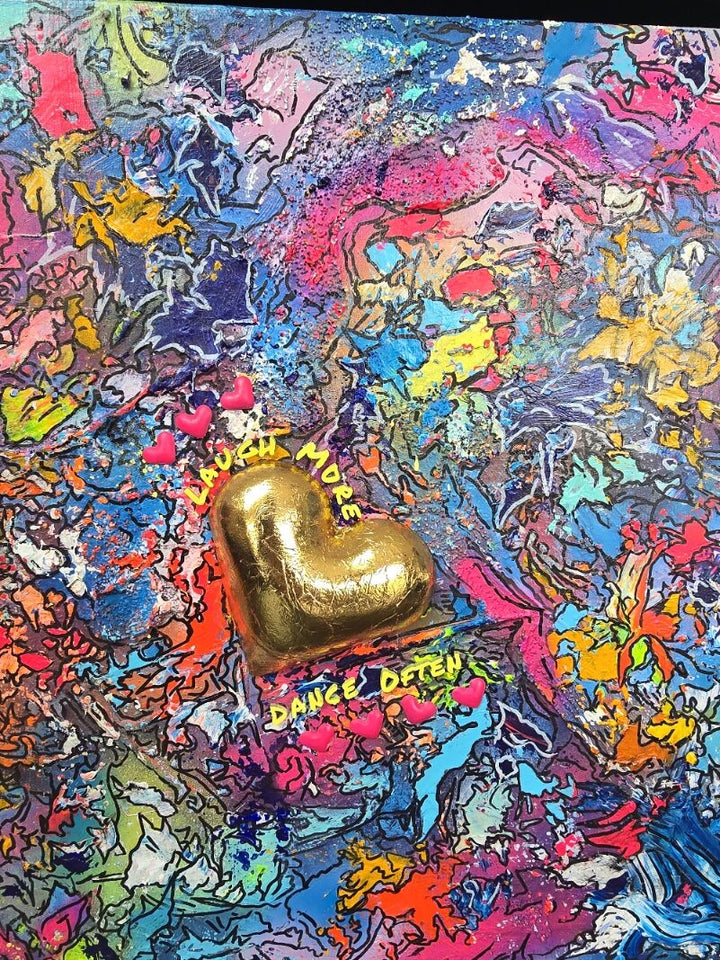 A vibrant abstract mixed-media painting bursting with swirling colours in pink, blue, yellow, orange and purple, with heavy texture and fluid forms. At the right side, a raised gold 3D heart sits prominently with text around it reading “Laugh More, Dance Often”. The artwork has an expressive, energetic surface with layered paint and resin details.