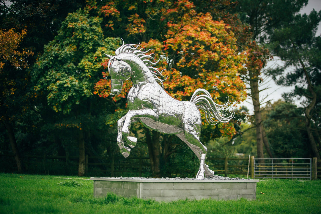 ‘Leaping Horse’ by Michael Turner, a unique, one-of-one stainless steel equestrian sculpture depicting a magnificent life-size horse leaping forward with its front legs raised and hind legs extended. Handcrafted from marine-grade stainless steel, the sculpture features intricately welded metallic plates, a powerful muscular body, flowing mane and sweeping tail. Its polished silver finish reflects the surrounding landscape, while the dynamic pose captures the strength, elegance and movement of a horse in ful