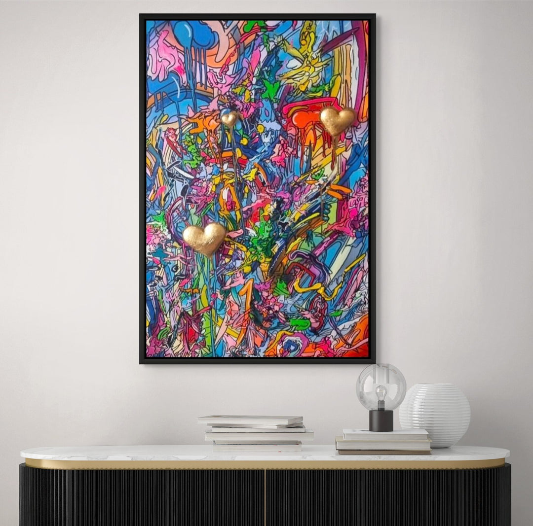 A vibrant mixed-media abstract artwork filled with layered graffiti-style lines, drips and splashes in bright pinks, blues, yellows and greens, featuring three raised gold 3D heart elements floating within the composition, framed in a black tray frame.