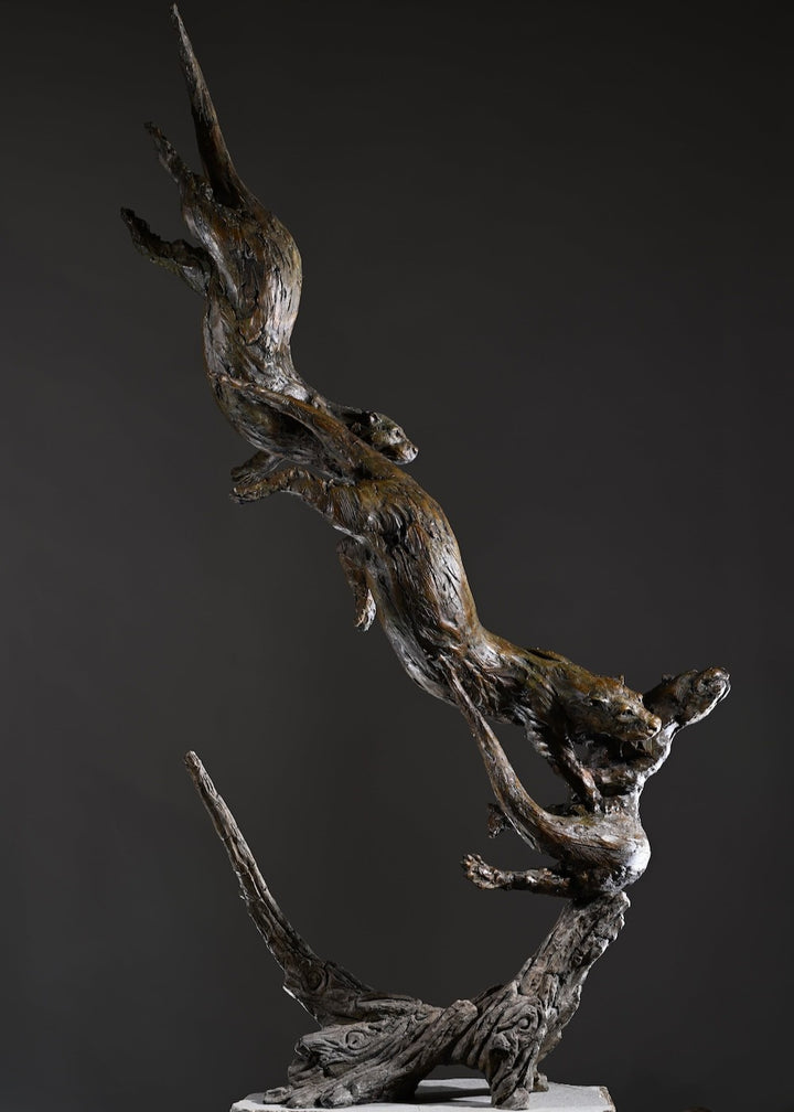 ‘Otters Playing Underwater’ by Hamish Mackie, a life-size limited-edition bronze wildlife sculpture depicting a mother otter and her playful cubs twisting, diving and swimming around a sculpted underwater tree root. The dynamic vertical composition features expressive animal movements, detailed fur textures and a weathered bronze finish in rich brown and earthy tones, capturing the playful energy and natural behaviour of otters beneath the water.