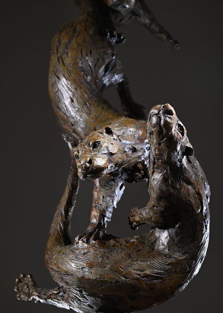‘Otters Playing Underwater’ by Hamish Mackie, a life-size limited-edition bronze wildlife sculpture depicting a mother otter and her playful cubs twisting, diving and swimming around a sculpted underwater tree root. The dynamic vertical composition features expressive animal movements, detailed fur textures and a weathered bronze finish in rich brown and earthy tones, capturing the playful energy and natural behaviour of otters beneath the water.