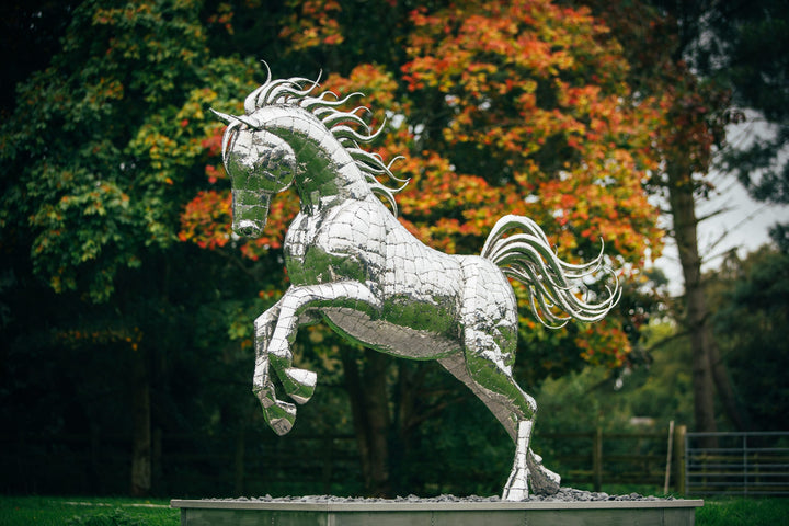 ‘Leaping Horse’ by Michael Turner, a unique, one-of-one stainless steel equestrian sculpture depicting a magnificent life-size horse leaping forward with its front legs raised and hind legs extended. Handcrafted from marine-grade stainless steel, the sculpture features intricately welded metallic plates, a powerful muscular body, flowing mane and sweeping tail. Its polished silver finish reflects the surrounding landscape, while the dynamic pose captures the strength, elegance and movement of a horse in ful