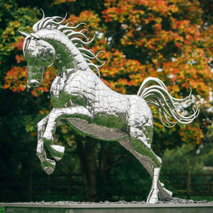 ‘Leaping Horse’ by Michael Turner, a unique, one-of-one stainless steel equestrian sculpture depicting a magnificent life-size horse leaping forward with its front legs raised and hind legs extended. Handcrafted from marine-grade stainless steel, the sculpture features intricately welded metallic plates, a powerful muscular body, flowing mane and sweeping tail. Its polished silver finish reflects the surrounding landscape, while the dynamic pose captures the strength, elegance and movement of a horse in ful